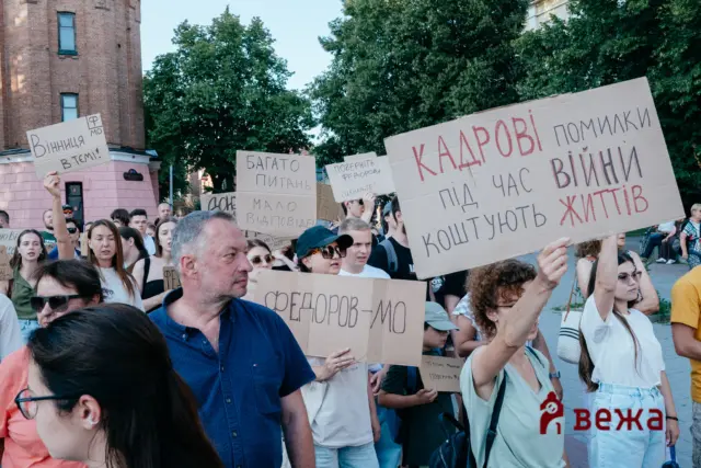 Crowd of protesters holding cardboard signs during a daytime demonstration in front of trees and a brick building.