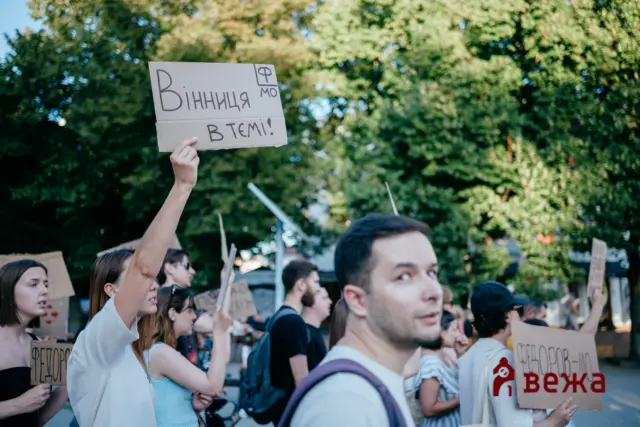 Protest crowd holding cardboard signs, foreground woman raising a sign with Cyrillic text in a park setting.