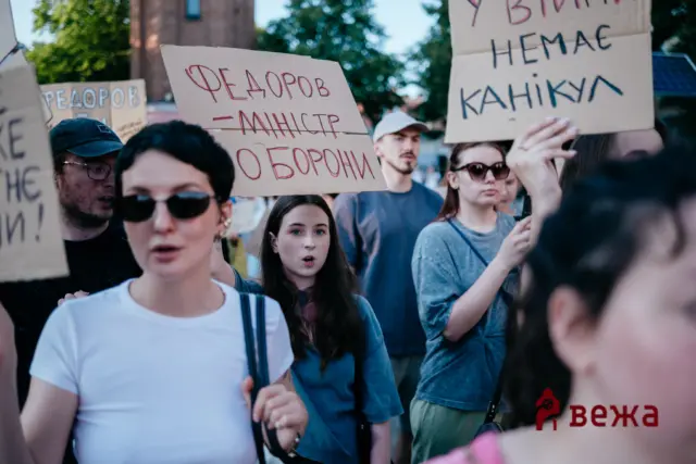 Protesters hold handmade cardboard signs during a daytime rally; one sign shows Cyrillic text.