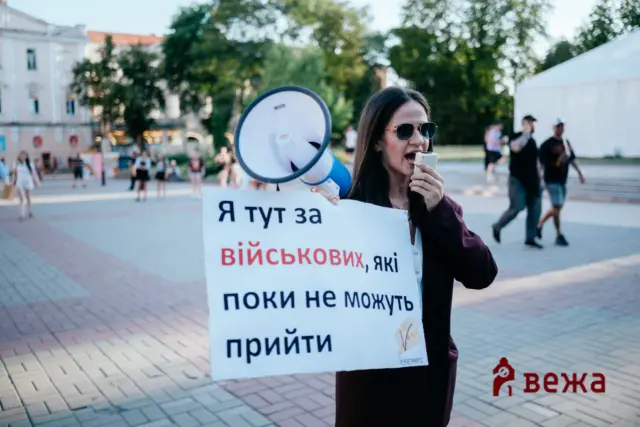 Protester woman with sunglasses using a megaphone, holding a Ukrainian protest sign in a public square.