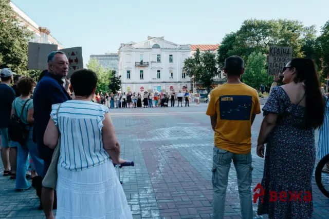 Crowd of people gathered in a city square holding protest signs, facing a white neoclassical building.