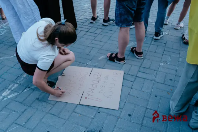 A person kneels on a paved surface writing red letters on cardboard while others stand nearby in casual clothing and sneakers.