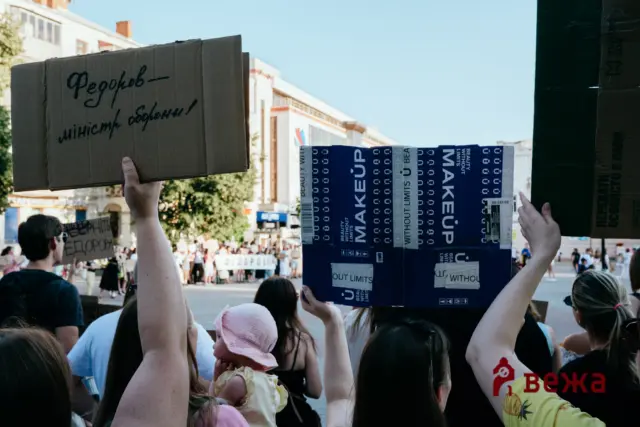 Protesters on a city street raise a large cardboard sign with handwritten Cyrillic text; background shows more demonstrators and tall buildings, plus blue boxes reading 'BEAUTY WITHOUT LIMITS'.